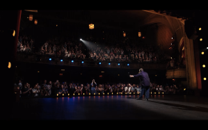 Wide shot from behind Patton Oswalt, on stage, arms outstretched at the end of his 2017 netflix comedy special, "Annihilation" as the crowd stands, arms also outstretched, upwards