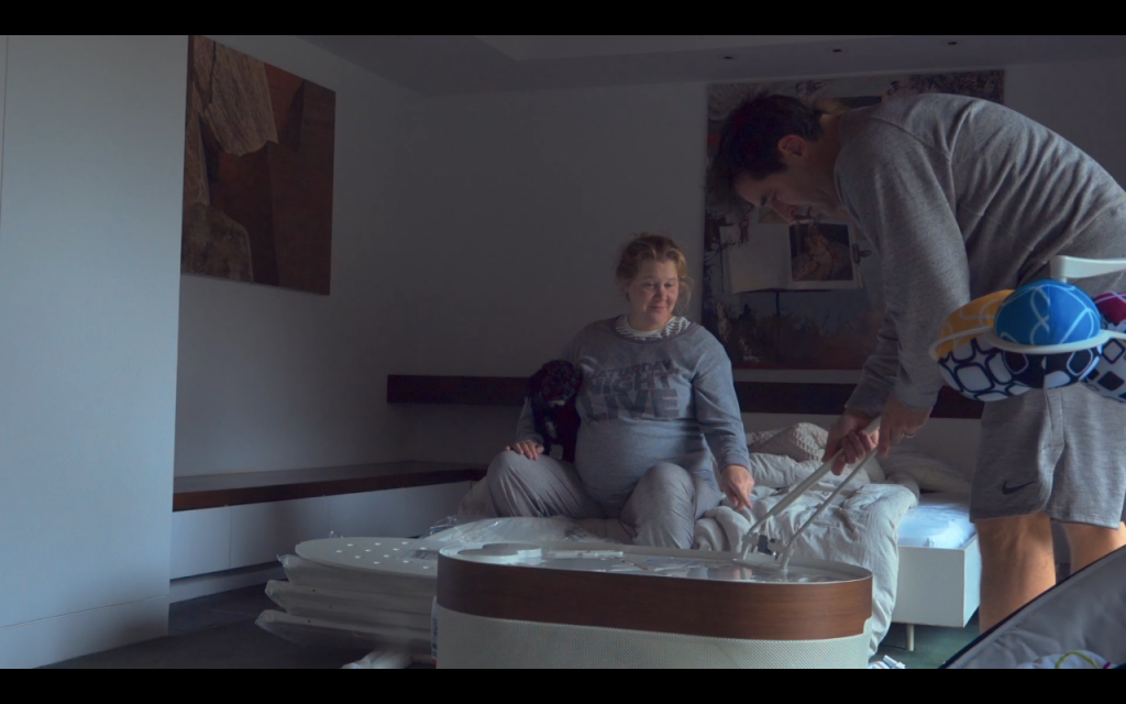 Amy Schumer sits in a Saturday Night Live grey sweatshirt and grey sweatpants on the bed holding dog while Chris Fisher also wears grey and puts together a crib in the HBO Max documentary Expecting Amy