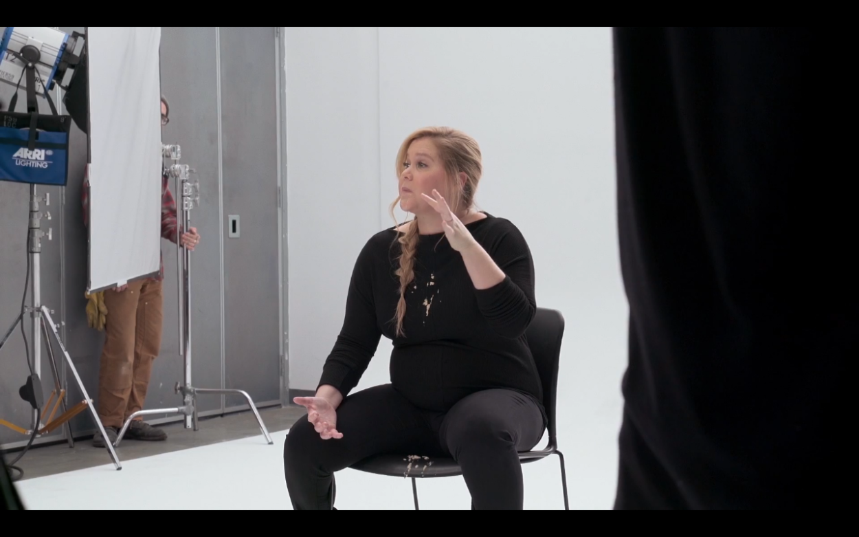 Amy Schumer sits down at a photo shoot for her 2019 special "Growing" wearing a black shirt and black pants, hopefully pretend puke on them. Background is white, with a pier 59 labelled Arri T2 in the background behind a diffusion