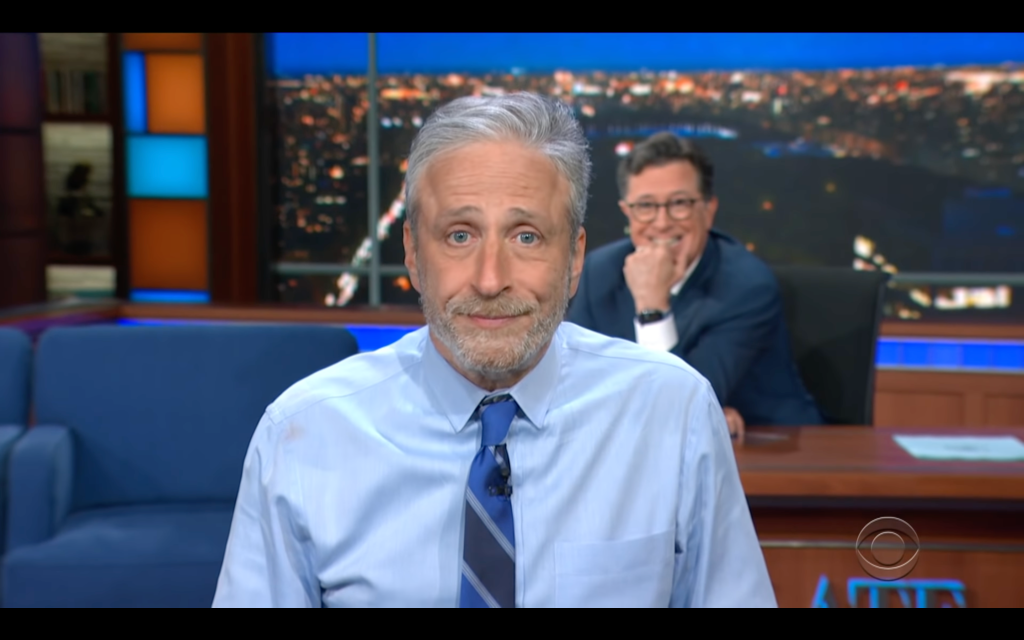 Jon Stewart appears on the Late Show with Stephen Colbert, June 14, 2021.  Jon has walked right up to the camera and is in focus, medium shot, wearing a blue button down shirt and blue striped tie, raised eyebrows and not quite smiling with wide eyes.  Stephen is laughing, sitting at his desk, elbow on desk fist raised to almost cover his mouth.
