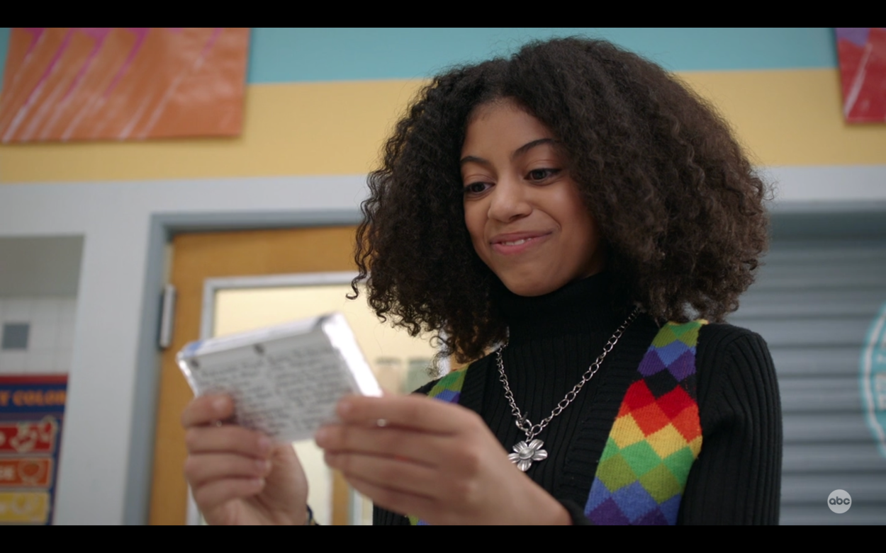 Arica Himmel smiles proudly looking at a mixtape in 2019's "Mixed-Ish". She wears a multicolored vest and black turtleneck with silver flower necklace.